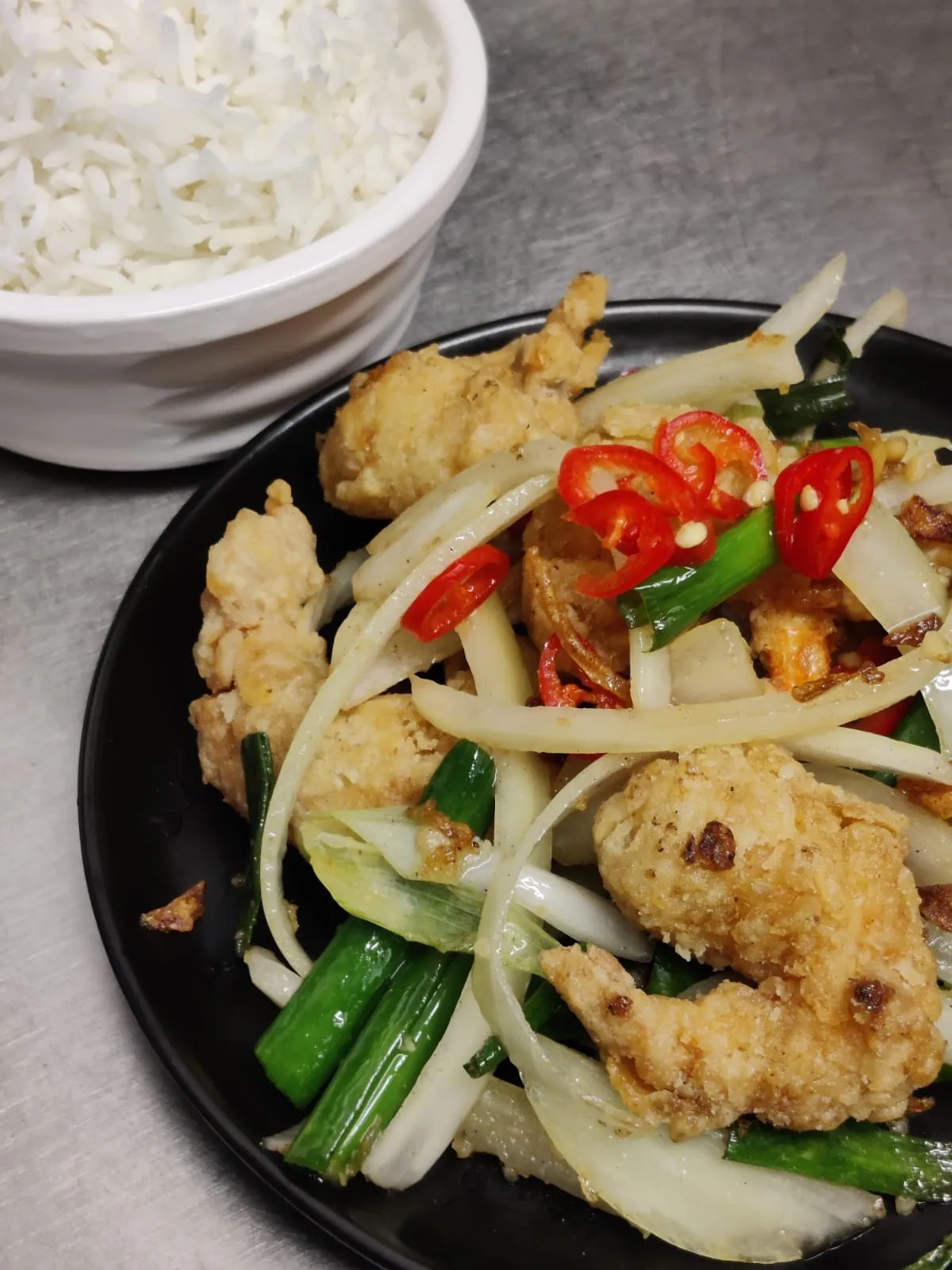 Light battered chicken with sliced onion, spring onion and fresh red chilli on a black plate, rice behind