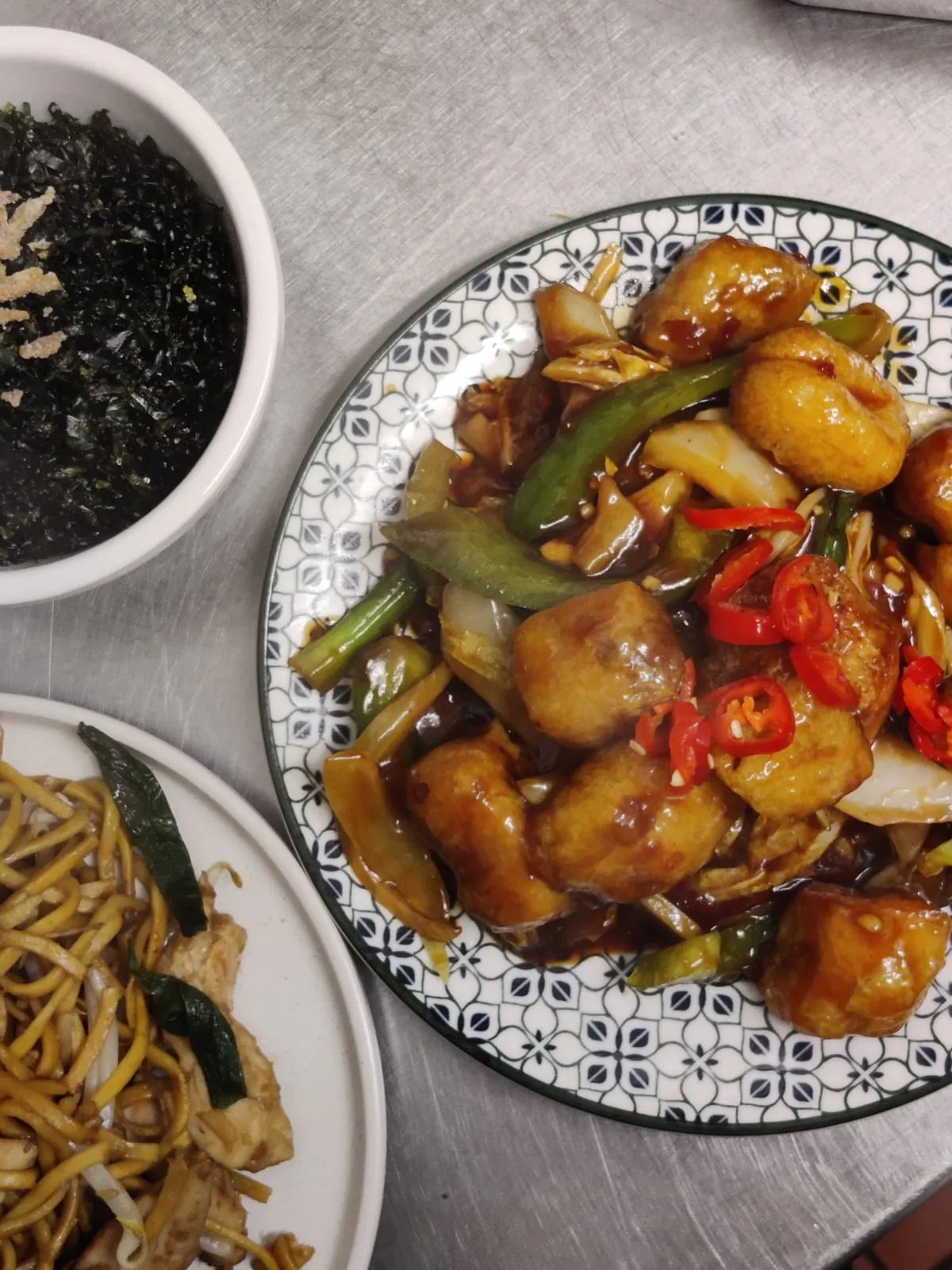 A stir-fry with green pepper, onion and chilli on a patterned plate, with crispy seaweed and noodles beside it