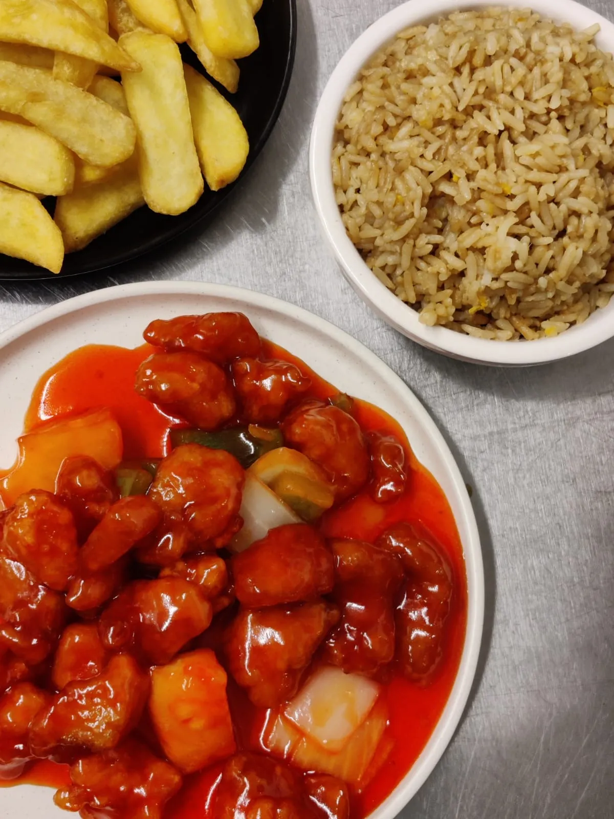 Battered pieces in glossy sweet and sour sauce with pineapple, beside chips and egg fried rice on a steel counter